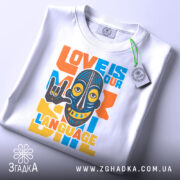 Футболка любов мова, біла, зображена зверху, фон світло-сірий, принт з написом 'LOVE IS OUR LANGUAGE' у яскравих кольорах, з малюнком маски, на футболці є етикетка з логотипом.