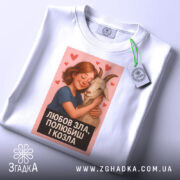Футболка любов зла, складена на столі, з білим кольором та яскравим принтом. На футболці зображена дівчина, яка обіймає козу, з сердечками навколо. На футболці є етикетка з логотипом, що підкреслює стиль.