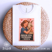 Футболка любов зла, складена на плетеному кошику, з білим кольором та яскравим принтом. Принт з дівчиною, яка обіймає козу, оточений сердечками, на фоні натурального матеріалу кошика, що додає текстури.