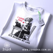 Футболка любовні історії. Біла футболка, складена на поверхні, з принтом дівчини за друкарською машинкою. На принті червоним написано 'I LOVE YOU', а нижче чорним 'IT'S RUINING MY LIFE'. На футболці є етикетка з зеленим шнурком. Фон світло-сірий.