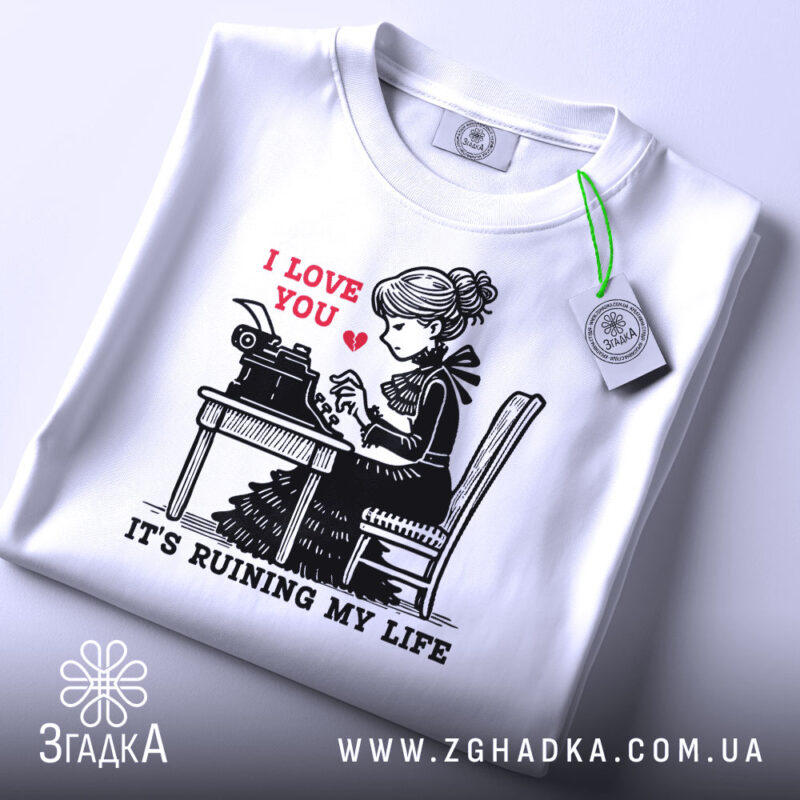 Футболка любовні історії. Біла футболка, складена на поверхні, з принтом дівчини за друкарською машинкою. На принті червоним написано 'I LOVE YOU', а нижче чорним 'IT'S RUINING MY LIFE'. На футболці є етикетка з зеленим шнурком. Фон світло-сірий.