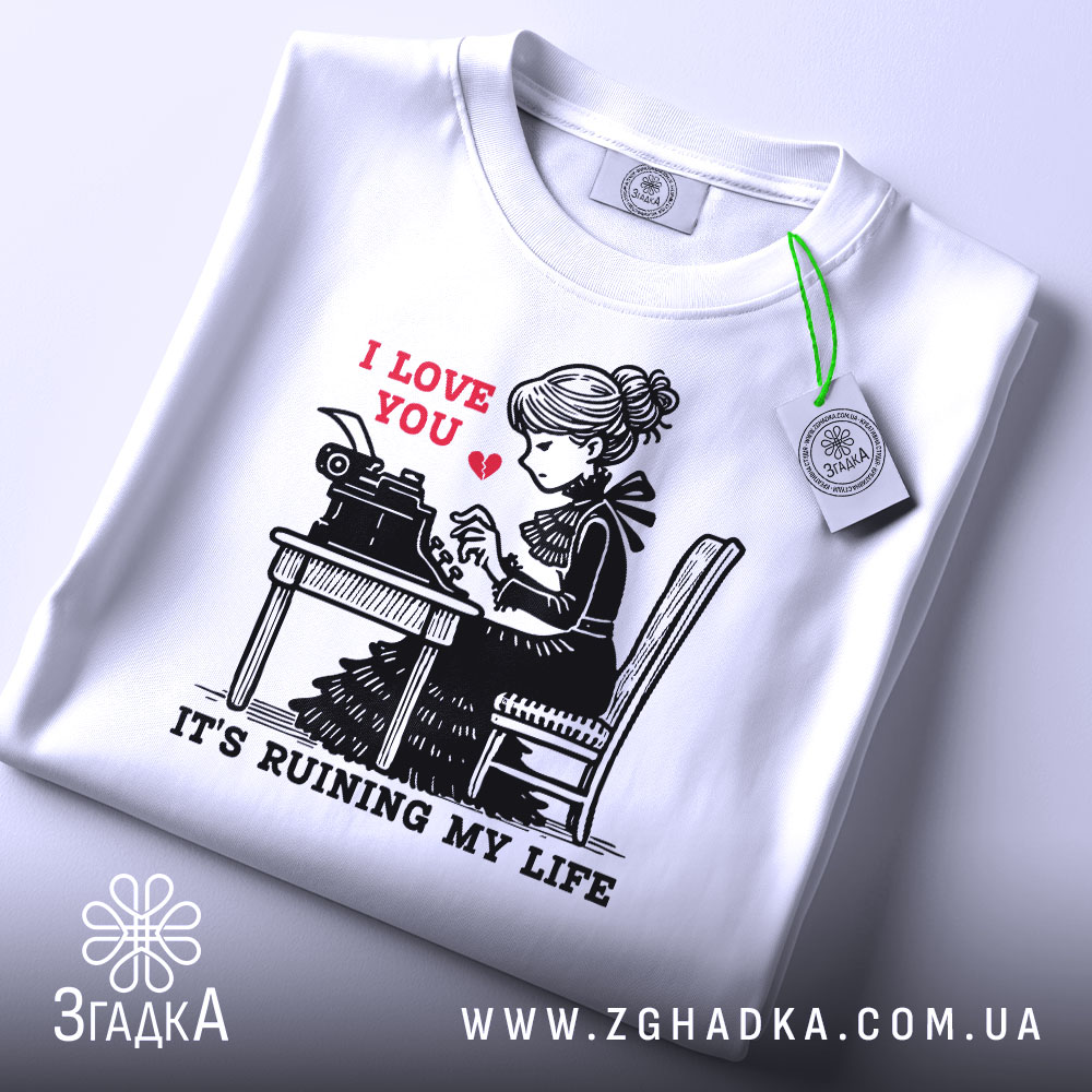 Футболка любовні історії. Біла футболка, складена на поверхні, з принтом дівчини за друкарською машинкою. На принті червоним написано 'I LOVE YOU', а нижче чорним 'IT'S RUINING MY LIFE'. На футболці є етикетка з зеленим шнурком. Фон світло-сірий.