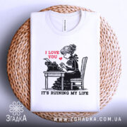 Футболка любовні історії. Біла футболка, складена на плетеній основі, з принтом дівчини за друкарською машинкою. На принті червоним написано 'I LOVE YOU', а нижче чорним 'IT'S RUINING MY LIFE'. Фон світло-сірий.