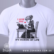 Футболка любовні історії. Біла футболка, складена на поверхні, з принтом дівчини за друкарською машинкою. На принті червоним написано 'I LOVE YOU', а нижче чорним 'IT'S RUINING MY LIFE'. На футболці є етикетка з зеленим шнурком. Фон світло-сірий.