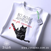 Футболка люди діляться: складена біла футболка з принтом чорного кота у окулярах, що тримає бензопилу з червоною плямою, на фоні світло-сірого кольору, з етикеткою зеленого кольору.