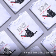 Футболка люди діляться: біла футболка на манекені, з принтом чорного кота у окулярах, що тримає бензопилу з червоною плямою, фон світло-сірий.