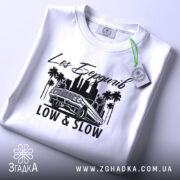 Футболка Лос Бердичів білого кольору, складена на поверхні, з принтом чорного кольору, що містить напис 'Los Бердичів' та малюнок автомобіля, з зеленим ярликом на комірці.