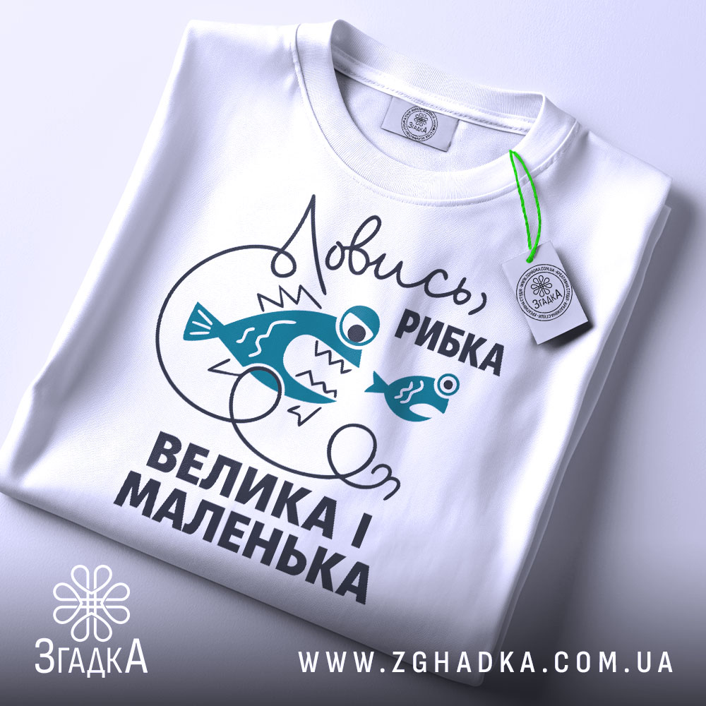 Футболка ловись рибка, біла, складена на поверхні, фон світло-сірий, принт з двома рибами та текстом "Ловись, рибка, велика і маленька" у темно-блакитному кольорі, з етикеткою на комірці.