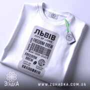 Футболка Львів місто історії, біла футболка, складена на столі, фон світлий, принт у вигляді етикетки з текстом 'FREEDOM BREW', прикріплений ярлик зеленого кольору.