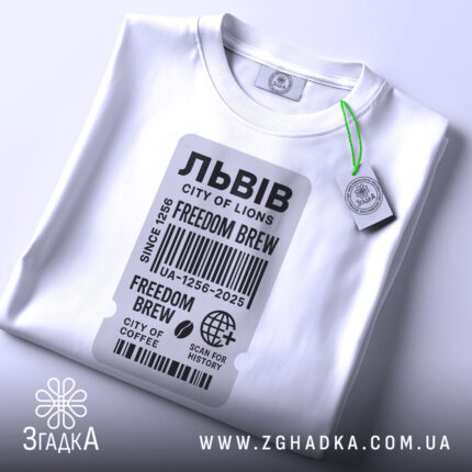 Футболка Львів місто історії, біла футболка, складена на столі, фон світлий, принт у вигляді етикетки з текстом 'FREEDOM BREW', прикріплений ярлик зеленого кольору.