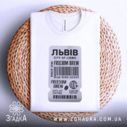 Футболка Львів місто історії, біла футболка, складена на плетеній кошику, фон світлий, принт у вигляді етикетки з текстом 'FREEDOM BREW' та штрих-кодом.