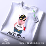 Футболка лихі 90, біла, складена на столі, фон світло-сірий, принт з мультяшним поросям у кепці та сонцезахисних окулярах, з написом 'МАФІЯ' на футболці, на комірці видна етикетка.