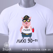 Футболка лихі 90, біла, складена на столі, фон світло-сірий, принт з мультяшним поросям у кепці та сонцезахисних окулярах, з написом 'МАФІЯ' на футболці, на комірці видна етикетка.