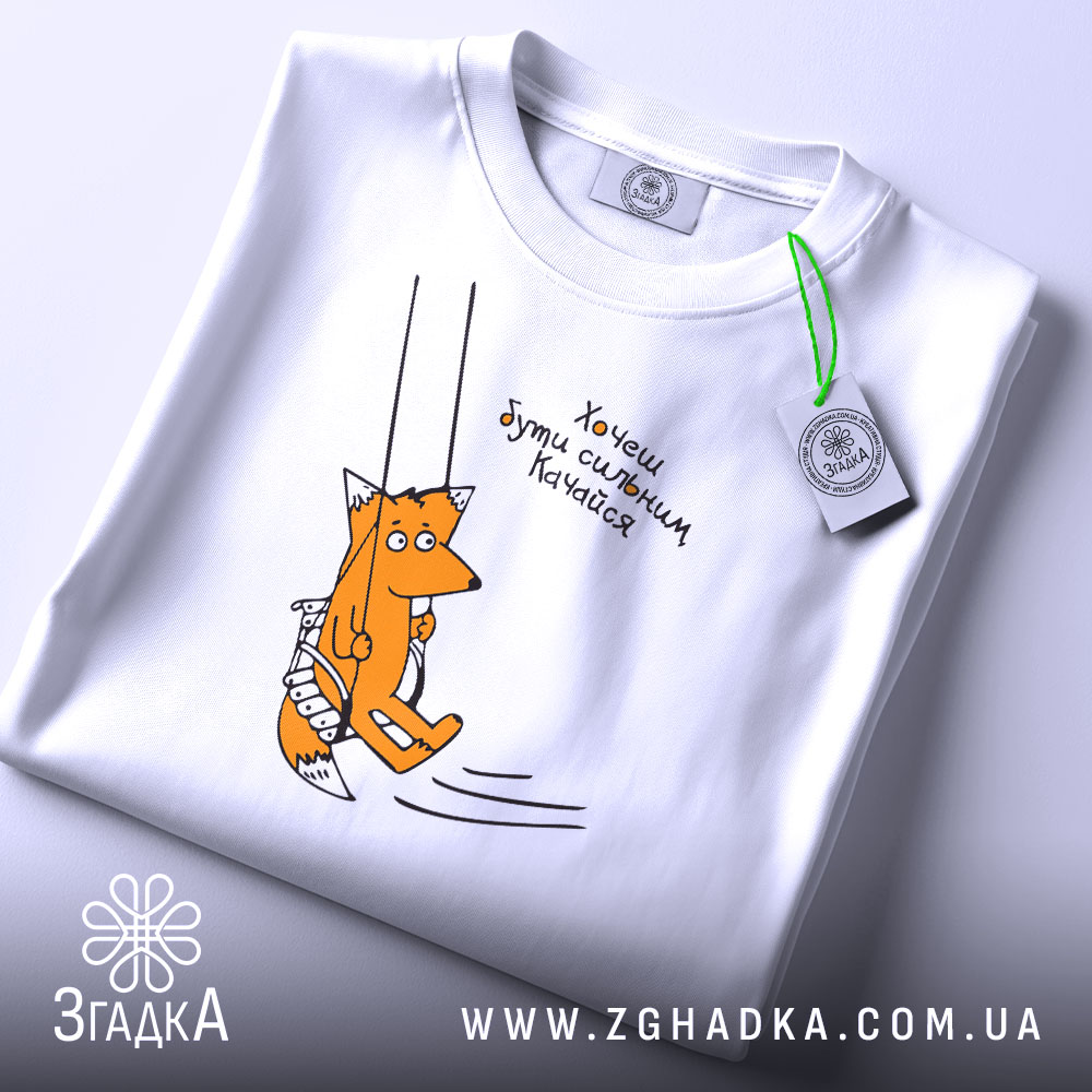 Футболка лисичка качайся, складена біла футболка на світлому фоні, принт з лисичкою на гойдалці, текст українською мовою, етикетка з зеленим шнурком.