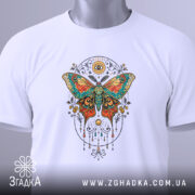 Футболка магія метелика білого кольору, складена на поверхні, зображена з верхнього ракурсу, фон світло-сірий, принт з метеликом в яскравих кольорах, на футболці є етикетка з зеленим шнурком.