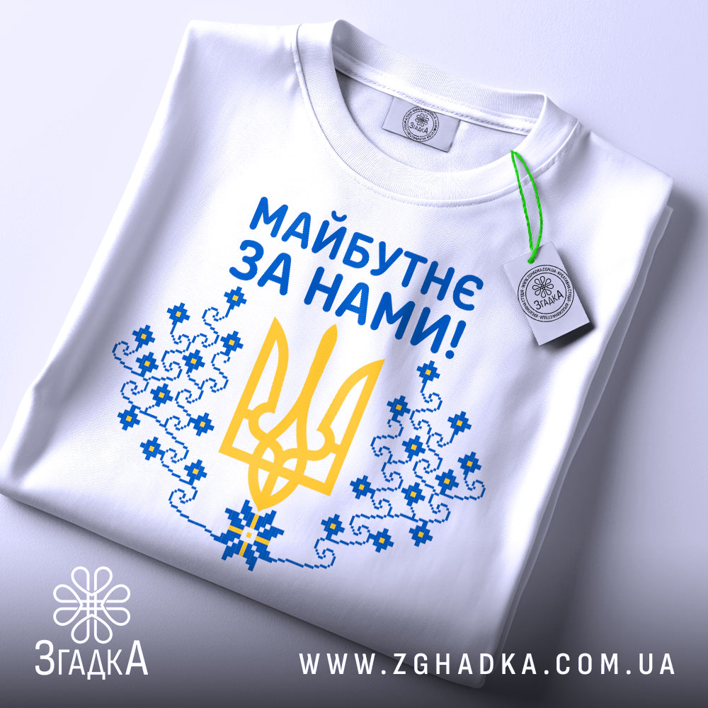 Футболка майбутнє за нами. Біла футболка, складена на столі, зображена зверху. Принт на футболці містить жовтий тризуб та сині квіти, текст "Майбутнє за нами!" написаний синім кольором, на футболці також є етикетка з зеленим шнурком.