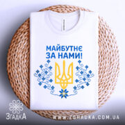 Футболка майбутнє за нами. Біла футболка, розкладена на плетеній підставці, зображена зверху. Принт на футболці містить жовтий тризуб та сині квіти, текст "Майбутнє за нами!" написаний синім кольором.