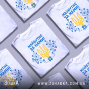 Футболка майбутнє за нами. Кілька білих футболок, складених на сірому фоні, зображені зверху. На всіх футболках присутній один і той же принт: жовтий тризуб та сині квіти, текст "Майбутнє за нами!" написаний синім кольором.