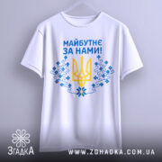 Футболка майбутнє за нами. Біла футболка, складена на столі, зображена зверху. Принт на футболці містить жовтий тризуб та сині квіти, текст "Майбутнє за нами!" написаний синім кольором, на футболці також є етикетка з зеленим шнурком.