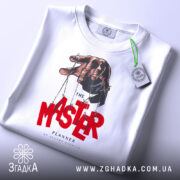 Футболка майстер планів, білого кольору, складена на поверхні, фон світло-сірий, принт з рукою, що тримає ляльку, червоний текст 'MASTER' та чорний 'PLANNER', з етикеткою зеленого кольору.