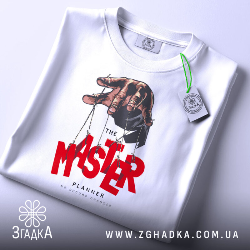 Футболка майстер планів, білого кольору, складена на поверхні, фон світло-сірий, принт з рукою, що тримає ляльку, червоний текст 'MASTER' та чорний 'PLANNER', з етикеткою зеленого кольору.