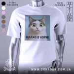 Футболка майже в нормі: біла футболка на манекені, з принтом кота з великими очима на блакитному фоні, текст під принтом чорний, фон світло-сірий.