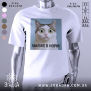 Футболка майже в нормі: біла футболка на манекені, з принтом кота з великими очима на блакитному фоні, текст під принтом чорний, фон світло-сірий.