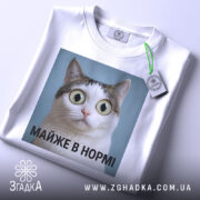 Футболка майже в нормі: складена біла футболка з принтом кота на блакитному фоні, текст під принтом чорний, на футболці є етикетка з зеленим шнурком.