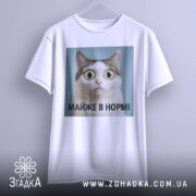 Футболка майже в нормі: біла футболка підвішена на вішалці, з принтом кота з великими очима на блакитному фоні, текст під принтом чорний, фон світло-сірий.