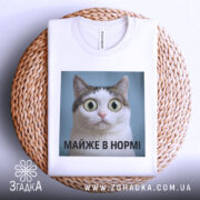 Футболка майже в нормі: складена біла футболка на плетеній основі, з принтом кота на блакитному фоні, текст під принтом чорний, фон світло-сірий.