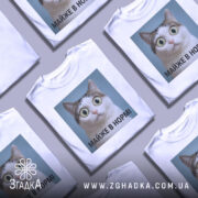 Футболка майже в нормі: біла футболка на манекені, з принтом кота з великими очима на блакитному фоні, текст під принтом чорний, фон світло-сірий.