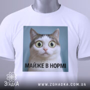 Футболка майже в нормі: складена біла футболка з принтом кота на блакитному фоні, текст під принтом чорний, на футболці є етикетка з зеленим шнурком.
