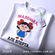 Футболка маленька вперта, білого кольору, зображена зверху, на ній видно етикетку, фон світло-сірий, принт з малюнком дівчинки в блакитній сукні з червоними бантиками на волоссі та написом 'МАЛЕНЬКА, АЛЕ ВПЕРТА.'