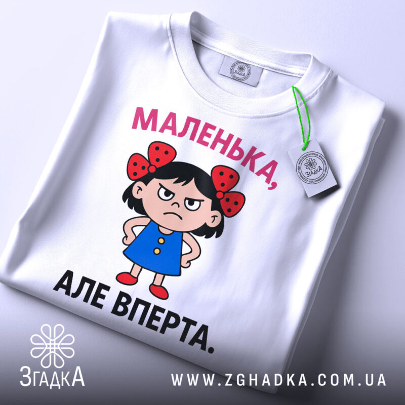 Футболка маленька вперта, білого кольору, зображена зверху, на ній видно етикетку, фон світло-сірий, принт з малюнком дівчинки в блакитній сукні з червоними бантиками на волоссі та написом 'МАЛЕНЬКА, АЛЕ ВПЕРТА.'