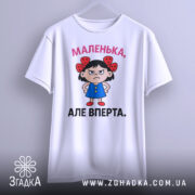 Футболка маленька вперта, білого кольору, підвішена на вішалці, фон світло-сірий, принт з малюнком дівчинки в блакитній сукні з червоними бантиками на волоссі та написом 'МАЛЕНЬКА, АЛЕ ВПЕРТА.'