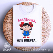 Футболка маленька вперта, білого кольору, зображена зверху на плетеній основі, фон світло-сірий, принт з малюнком дівчинки в блакитній сукні з червоними бантиками на волоссі та написом 'МАЛЕНЬКА, АЛЕ ВПЕРТА.'