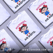 Футболка маленька вперта, білого кольору, зображена на манекені з правого ракурсу, фон світло-сірий, принт з малюнком дівчинки в блакитній сукні з червоними бантиками на волоссі та написом 'МАЛЕНЬКА, АЛЕ ВПЕРТА.'