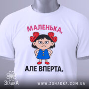 Футболка маленька вперта, білого кольору, зображена зверху, на ній видно етикетку, фон світло-сірий, принт з малюнком дівчинки в блакитній сукні з червоними бантиками на волоссі та написом 'МАЛЕНЬКА, АЛЕ ВПЕРТА.'