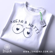 Футболка маленькі радості, біла, складена на столі, фон світло-сірий, принт чорного кольору з написом 'ЛЮБЛЮ В ЖИТТІ' та малюнком у вигляді сердець, на комірці є етикетка.