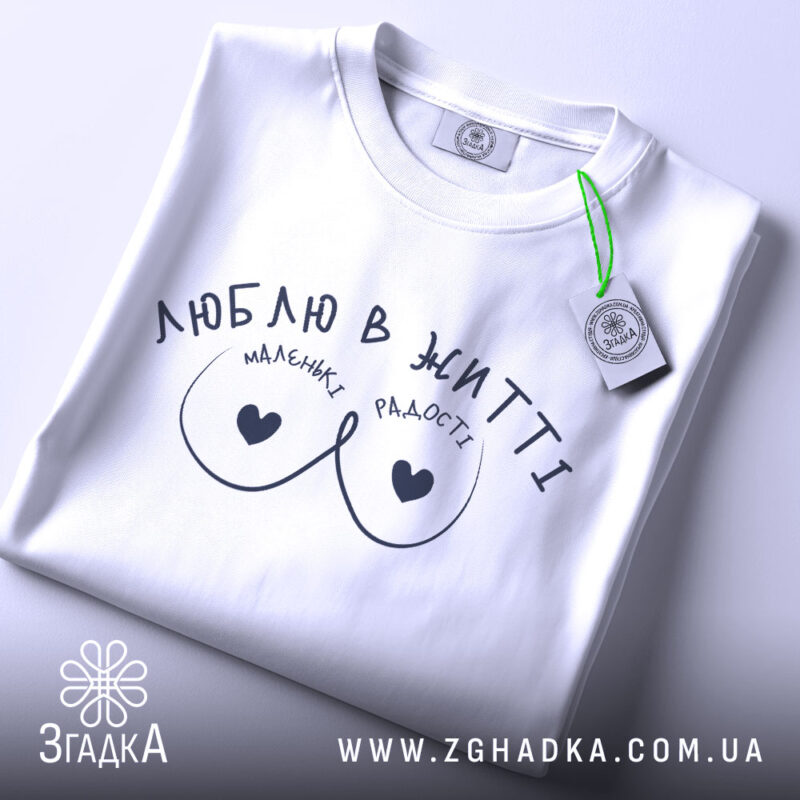 Футболка маленькі радості, біла, складена на столі, фон світло-сірий, принт чорного кольору з написом 'ЛЮБЛЮ В ЖИТТІ' та малюнком у вигляді сердець, на комірці є етикетка.