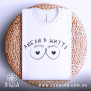 Футболка маленькі радості, біла, складена на плетеній кошику, фон світлий, принт чорного кольору з написом 'ЛЮБЛЮ В ЖИТТІ' та малюнком у вигляді сердець.