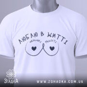 Футболка маленькі радості, біла, складена на столі, фон світло-сірий, принт чорного кольору з написом 'ЛЮБЛЮ В ЖИТТІ' та малюнком у вигляді сердець, на комірці є етикетка.