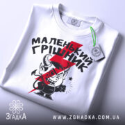 Футболка маленький грішник, біла, складена на поверхні, фон світло-сірий, принт з червоним і чорним малюнком маленького диявола та написом, з зеленим ярликом.