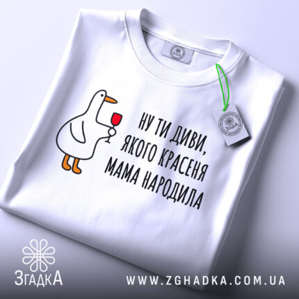 Футболка мама красеня народила, біла, складена на столі, фон світло-сірий, принт з качкою, що тримає келих, текст чорний, на етикетці зелена стрічка.