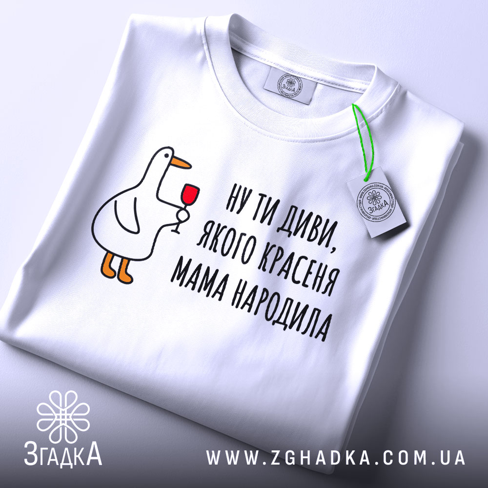 Футболка мама красеня народила, біла, складена на столі, фон світло-сірий, принт з качкою, що тримає келих, текст чорний, на етикетці зелена стрічка.