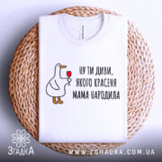 Футболка мама красеня народила, біла, складена на плетеній основі, фон світлий, принт з качкою, що тримає келих, текст чорний.