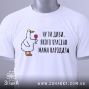 Футболка мама красеня народила, біла, складена на столі, фон світло-сірий, принт з качкою, що тримає келих, текст чорний, на етикетці зелена стрічка.