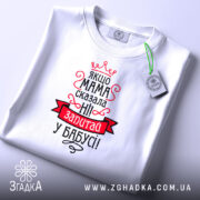 Футболка мама сказала ні. Біла футболка, складена на столі, з видимим принтом. Зображена з верхнього ракурсу. На фоні білий колір. Принт червоного та чорного кольорів з текстом та короною, з біркою на комірці.