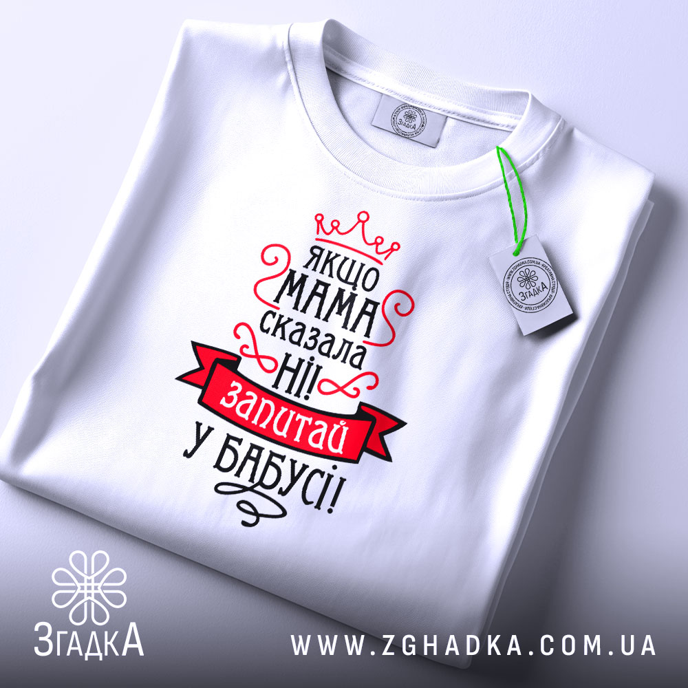 Футболка мама сказала ні. Біла футболка, складена на столі, з видимим принтом. Зображена з верхнього ракурсу. На фоні білий колір. Принт червоного та чорного кольорів з текстом та короною, з біркою на комірці.