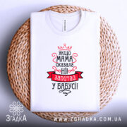 Футболка мама сказала ні. Біла футболка, складена на плетеній основі, з видимим принтом. Зображена з верхнього ракурсу. На фоні світлий колір. Принт червоного та чорного кольорів з текстом та короною.